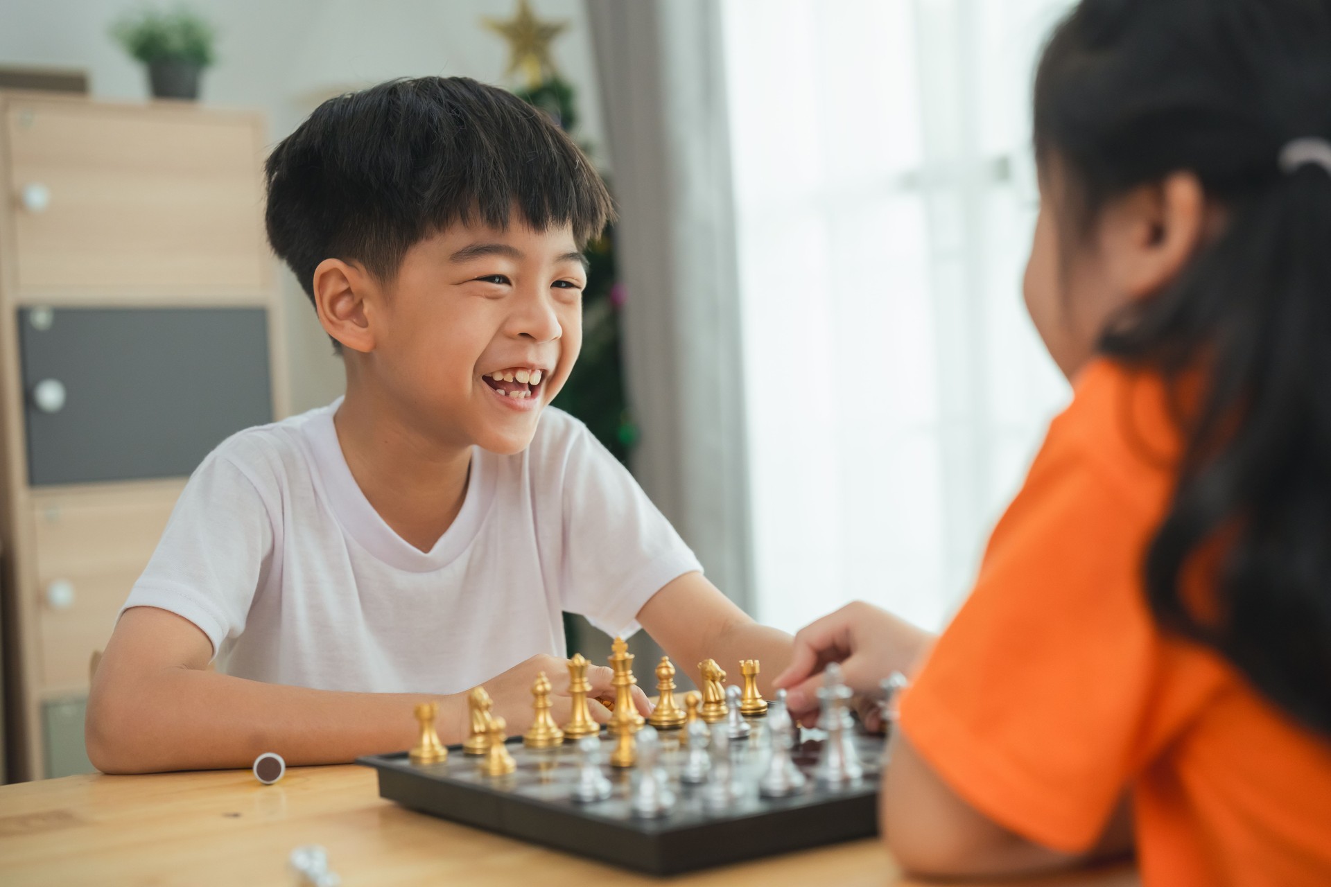 Two children playing a game of chess. One is smiling and the other is frowning. The boy in the white shirt is smiling and the girl in the orange shirt is frowning